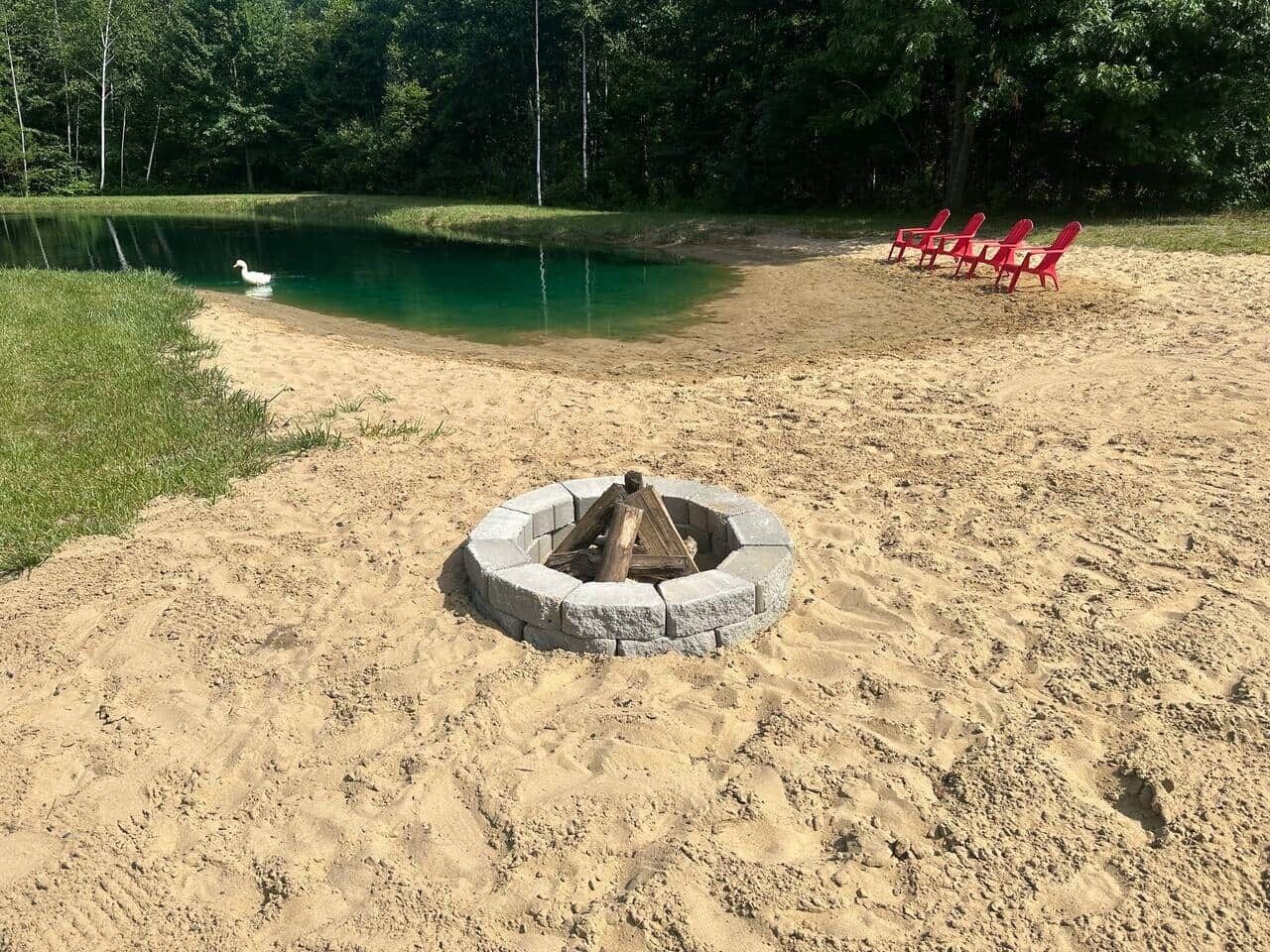 A sandy beach with a fire pit in the center, surrounded by red chairs, and a pond with a duck in the background.