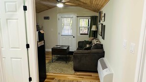 A living room with a wooden ceiling, a sofa, a coffee table, a window with curtains, and a door. - Hidden Oaks | Lodge Cabin | Unit 3 (Birch Run Township)