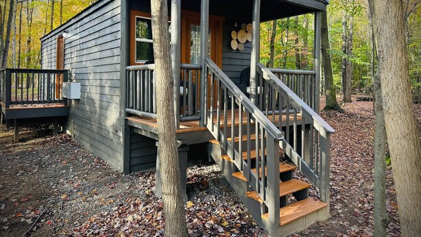 A cabin with a wooden deck, a porch, and a staircase leading to a covered entryway. - Hidden Oaks | Lodge Cabin | Unit 3 (Birch Run Township)