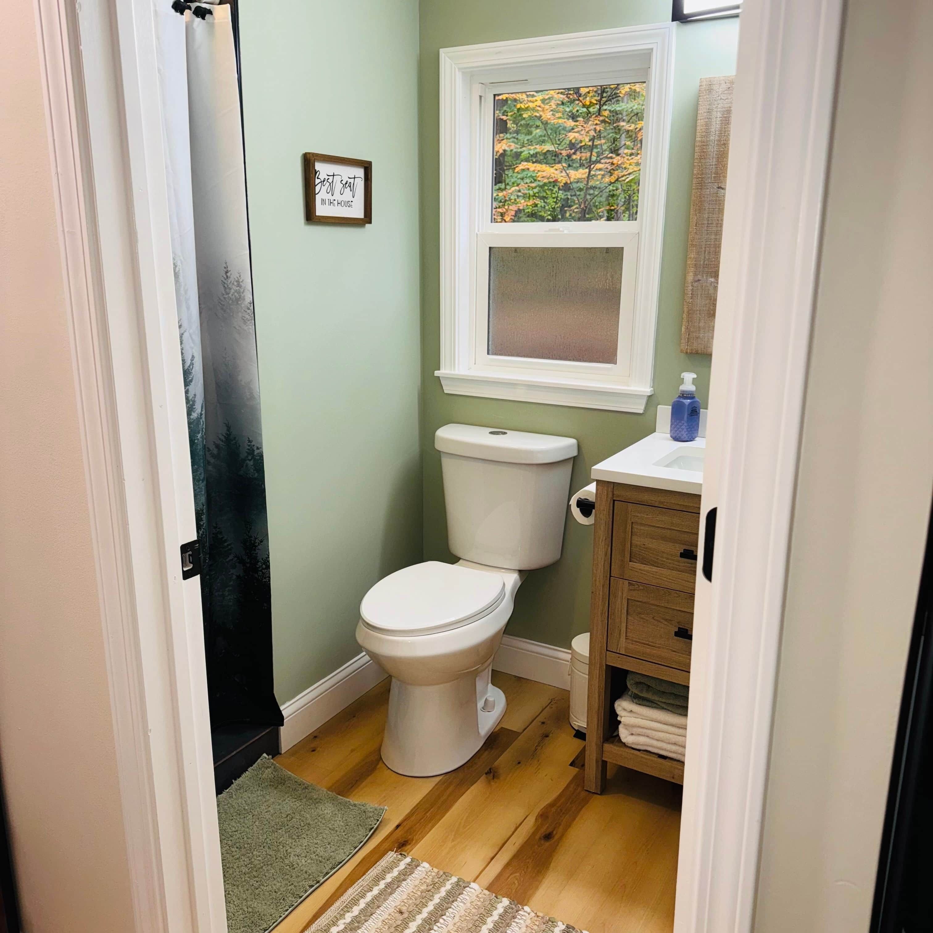 Une petite salle de bain avec des toilettes, une fenêtre donnant sur des arbres et un meuble en bois contenant des serviettes.