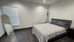 Un dormitorio con cama, dos ventanas con persianas, una lámpara de pie y un aparador de color oscuro.