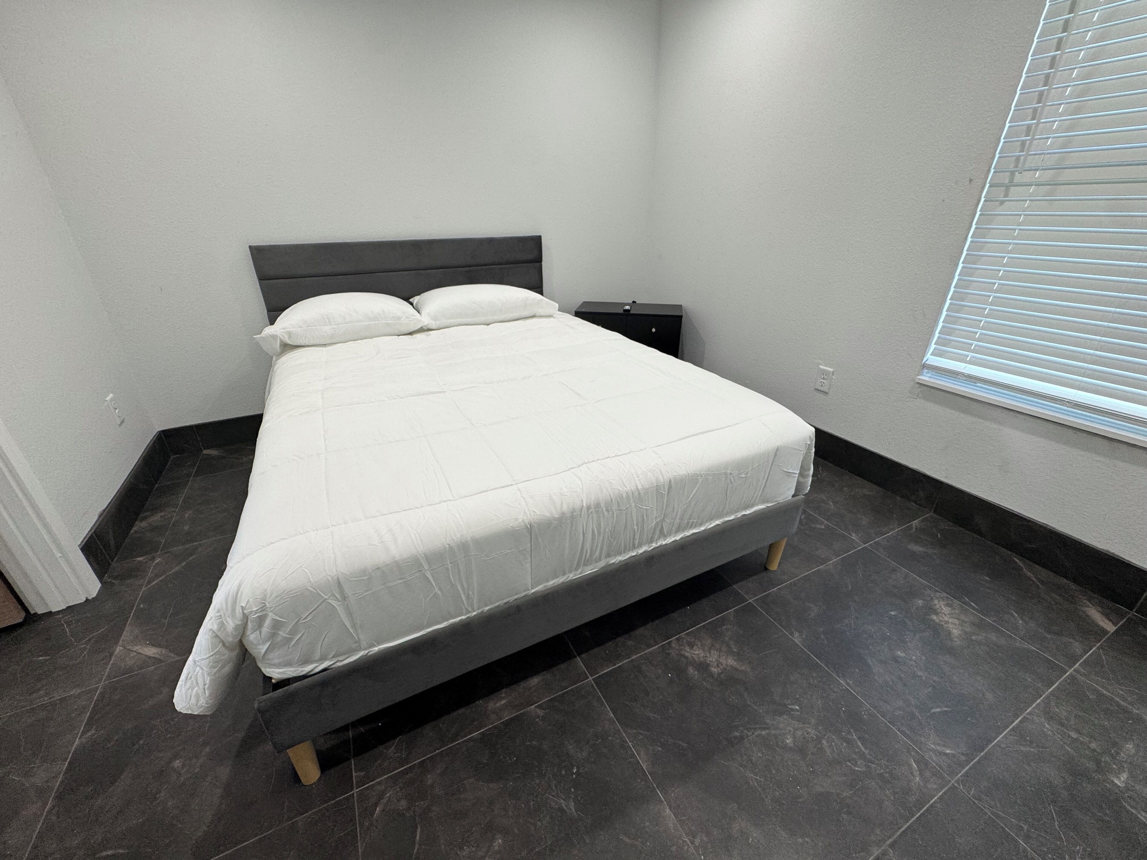 A neatly made bed in a room with dark tiled flooring and a simple headboard.