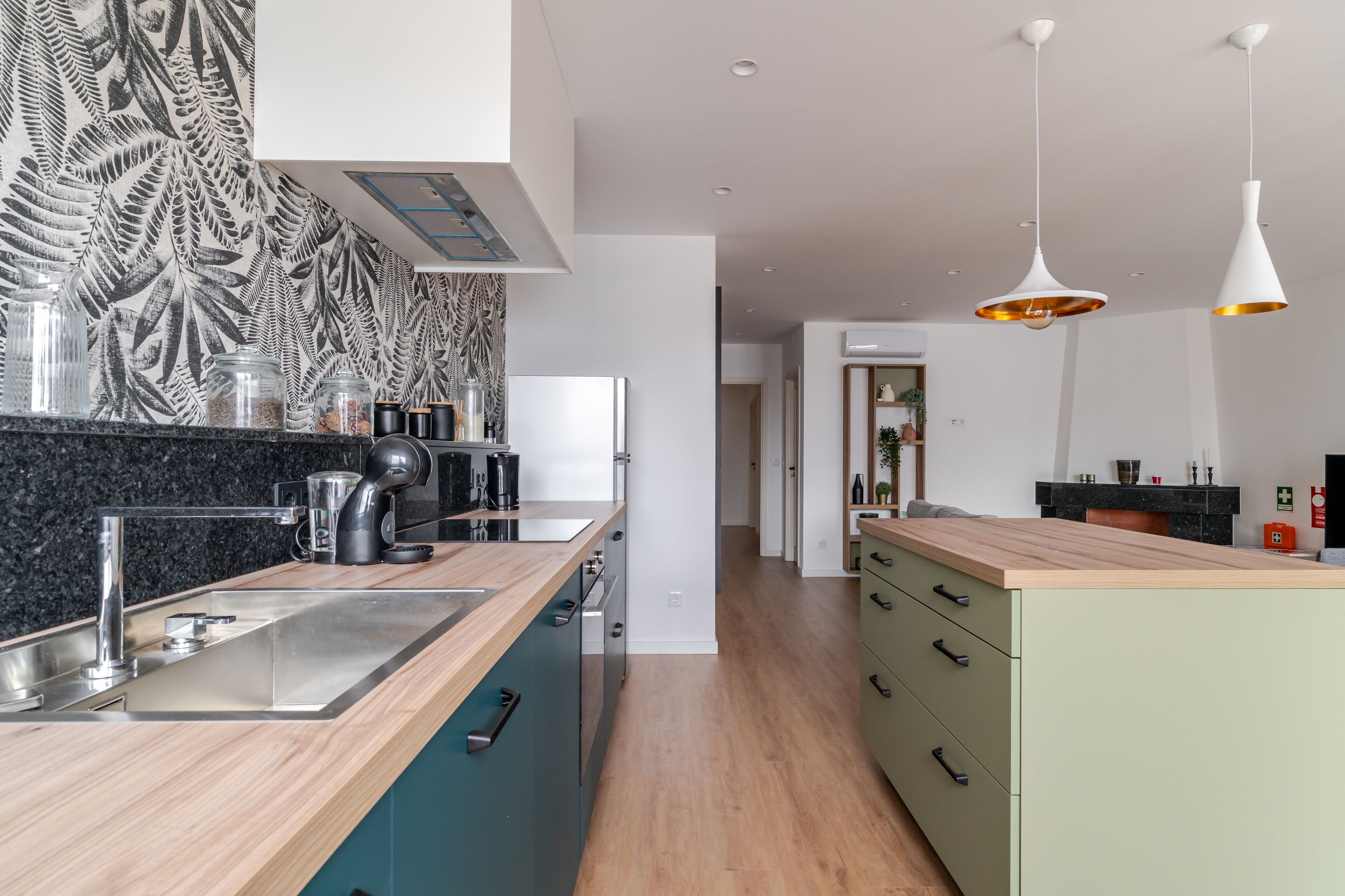 A modern kitchen with a wooden countertop, stainless steel sink, and a built-in oven. There are hanging pendant lights and a patterned backsplash.