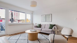 Un salon moderne avec un canapé, une table basse ronde et un fauteuil. Deux œuvres d’art encadrées ornent le mur, et une fenêtre offre une vue sur des immeubles et des arbres.