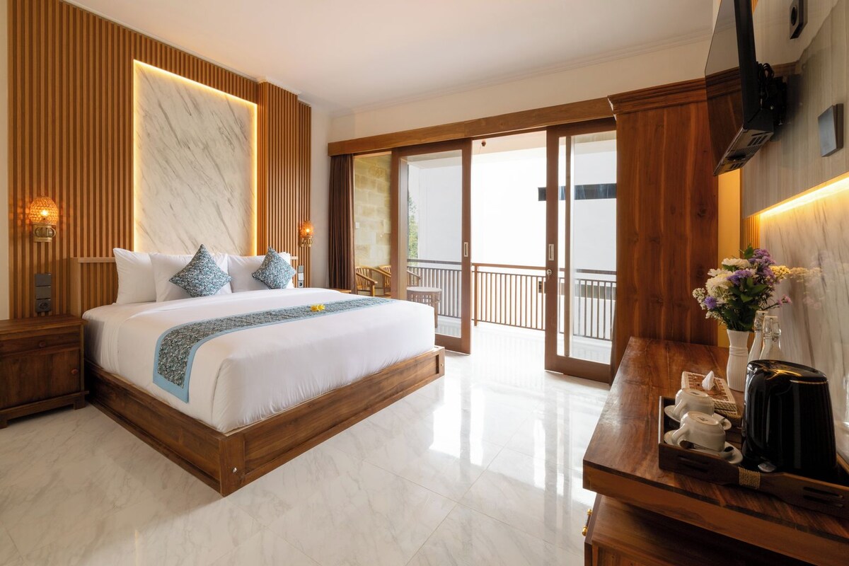 A hotel room with a large bed, wooden headboard, a wooden nightstand, a bedside table with a coffee maker, and a balcony with a view.