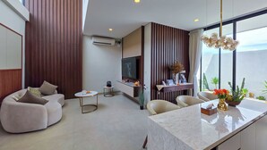 A modern living room with a sofa, a coffee table, and a TV. There is a kitchen area with a dining table and chairs. The room has a large window and a ceiling light. - Villa Sorano Seminyak by Nagisa Bali (Seminyak)