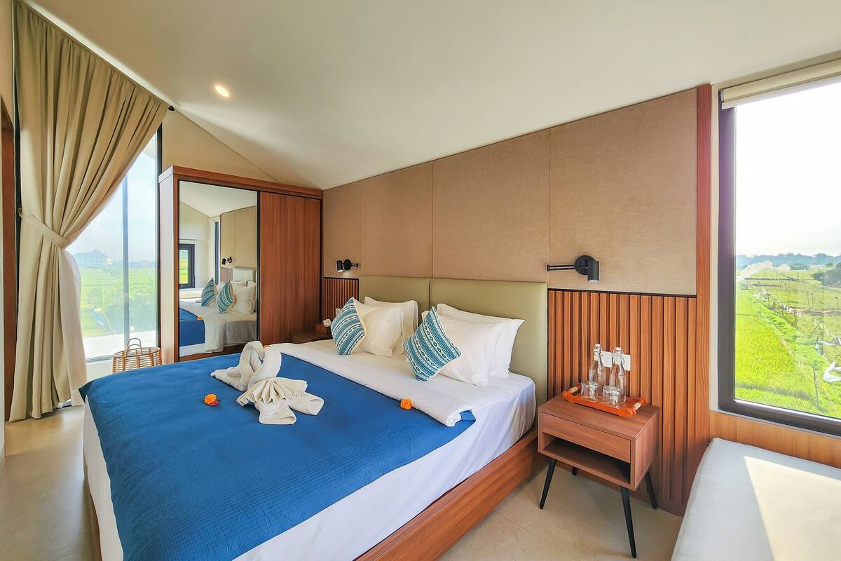 Une chambre d’hôtel avec un grand lit, une table de chevet, un miroir et une vue sur un paysage verdoyant.