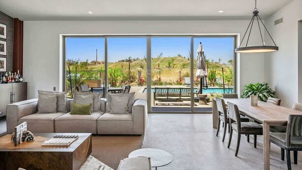 A modern living room with a large sliding glass door leading to a pool area, a dining table with chairs, and a sofa with cushions.