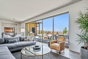 Un salón moderno con sofá, sillón y mesa de centro, ventanas amplias y vistas al exterior.