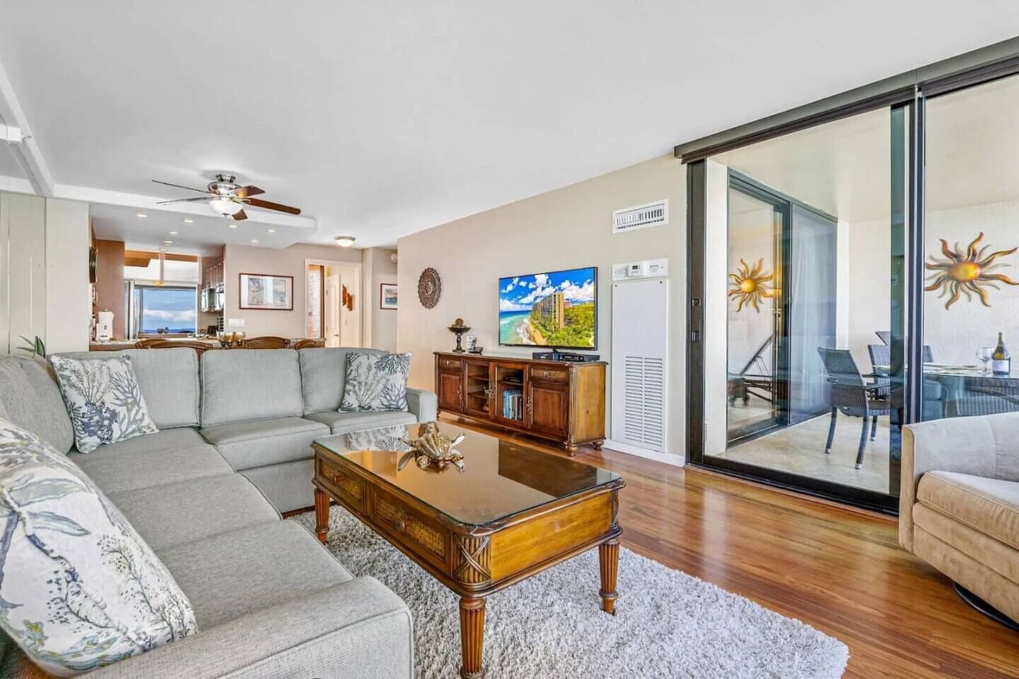 Ein großzügiges Wohnzimmer mit einem grauen Ecksofa, einem Holztisch und einem Fernseher. Eine Glastür führt auf einen Balkon mit Sitzgelegenheiten.