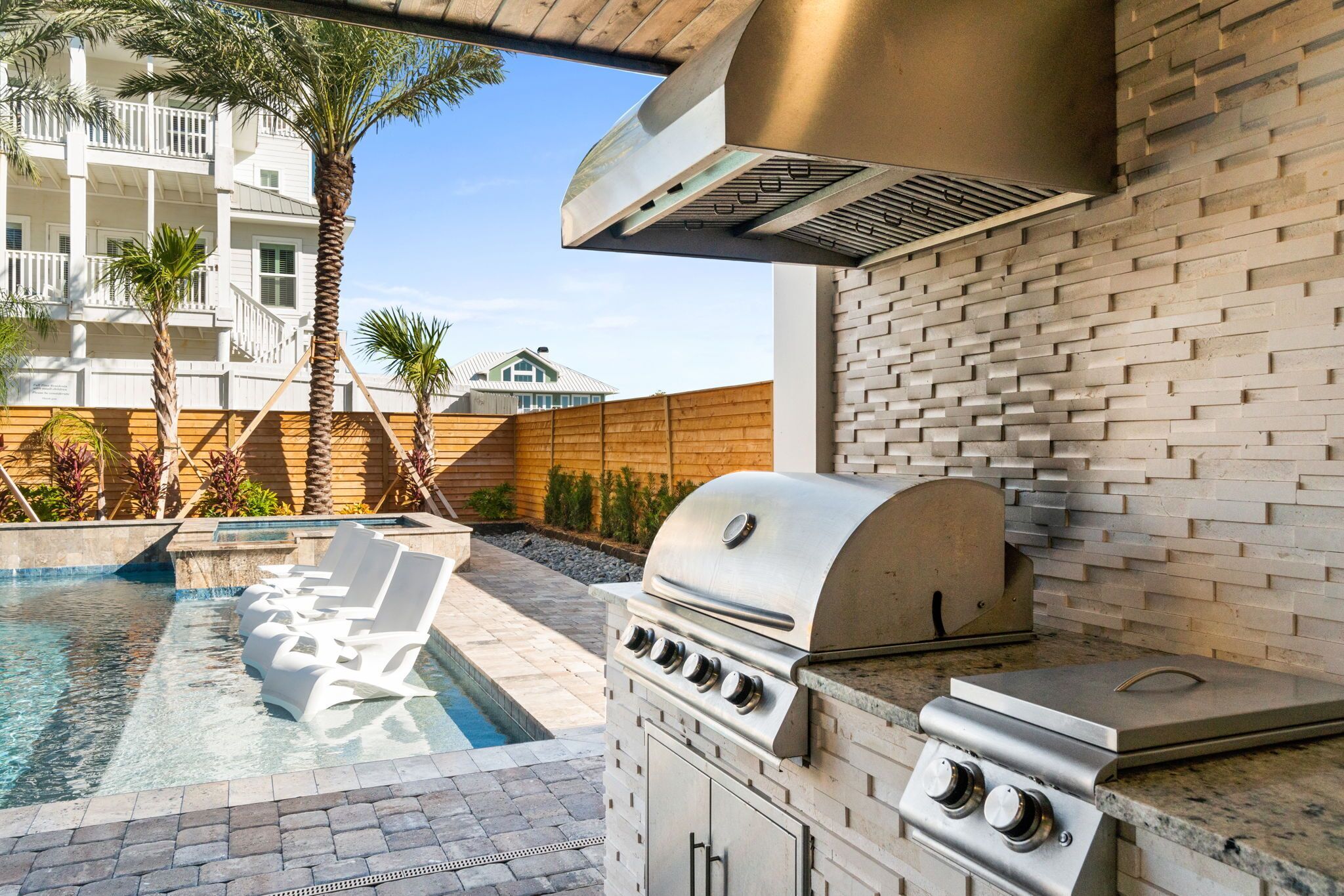 An outdoor kitchen with a grill, a pool with lounge chairs, and a wooden fence.