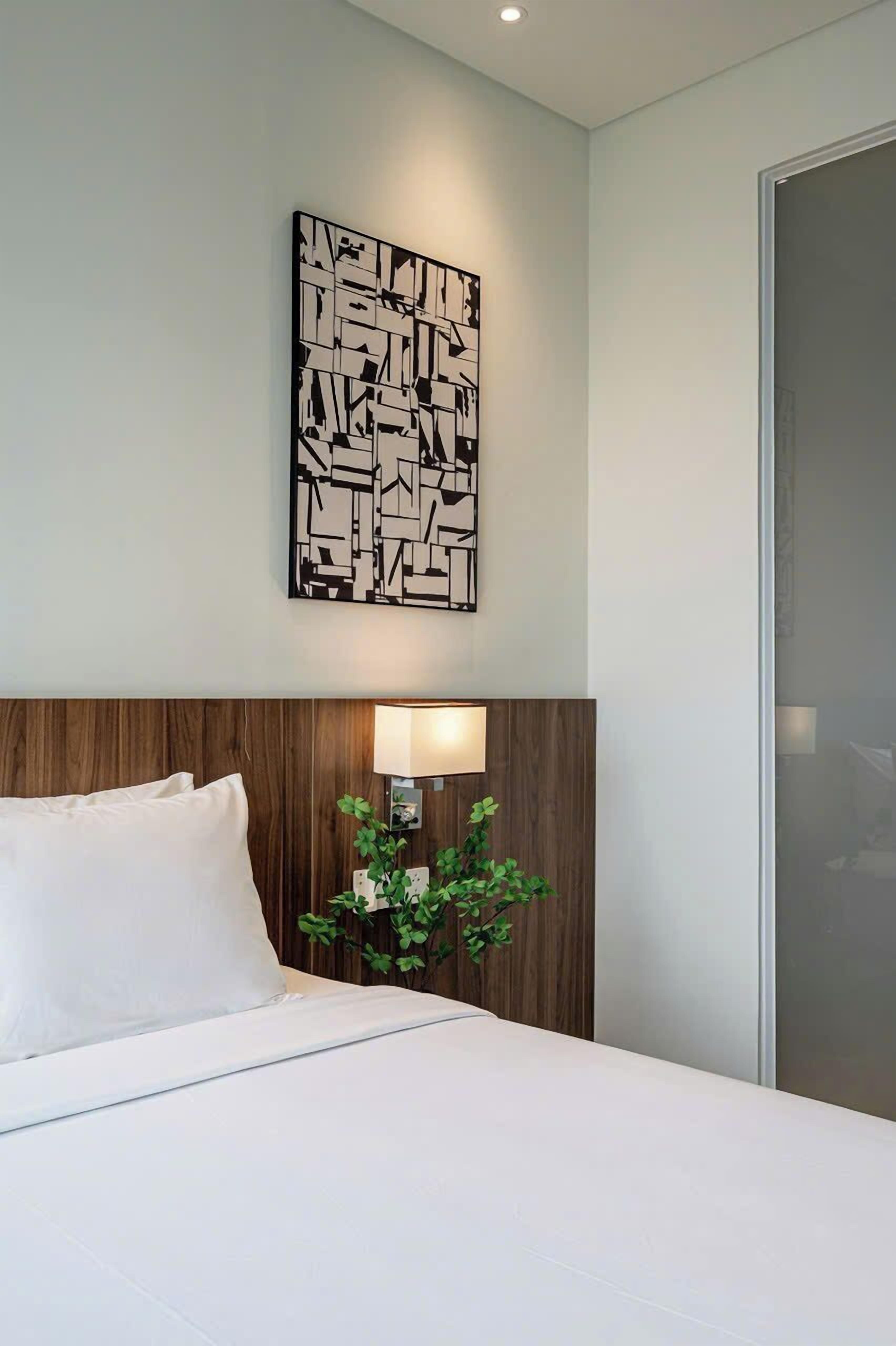 A hotel room with a bed, a wall-mounted abstract art piece, a bedside lamp, and a plant.
