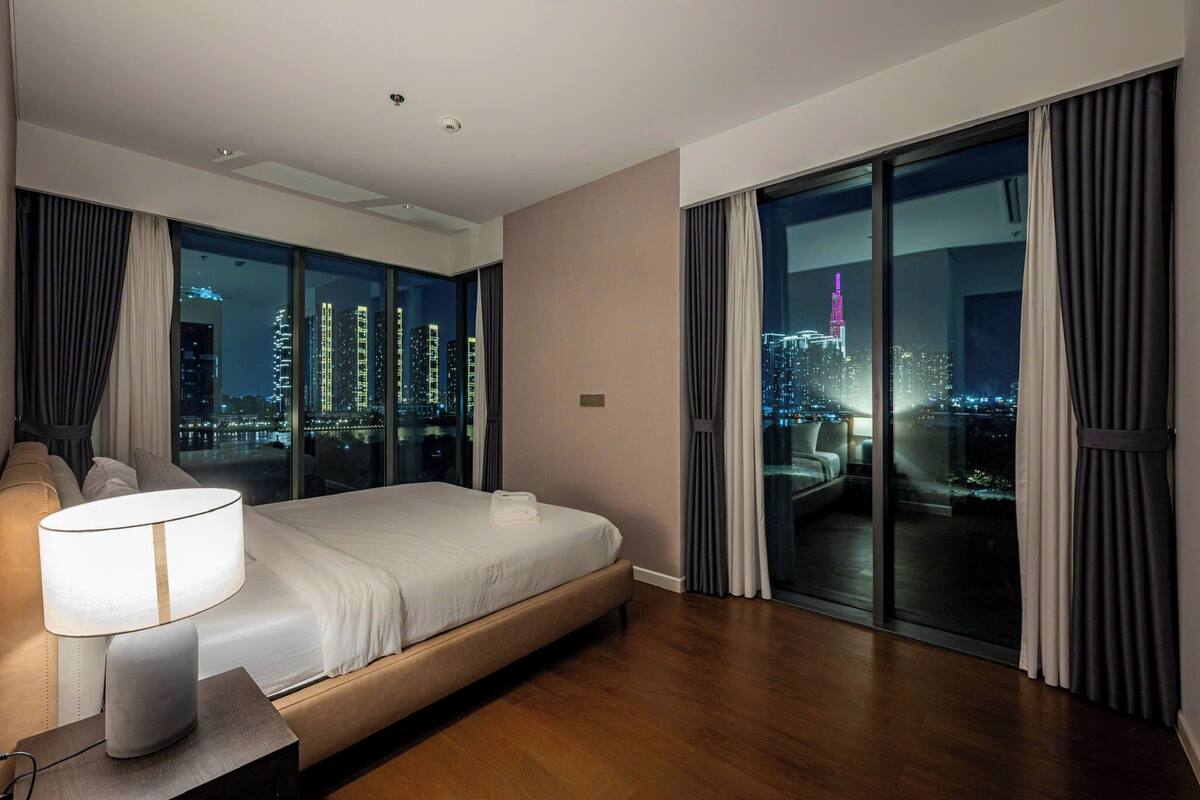 A modern hotel room with a large bed, a bedside table with a lamp, a city view through floor-to-ceiling windows, and a balcony with curtains.