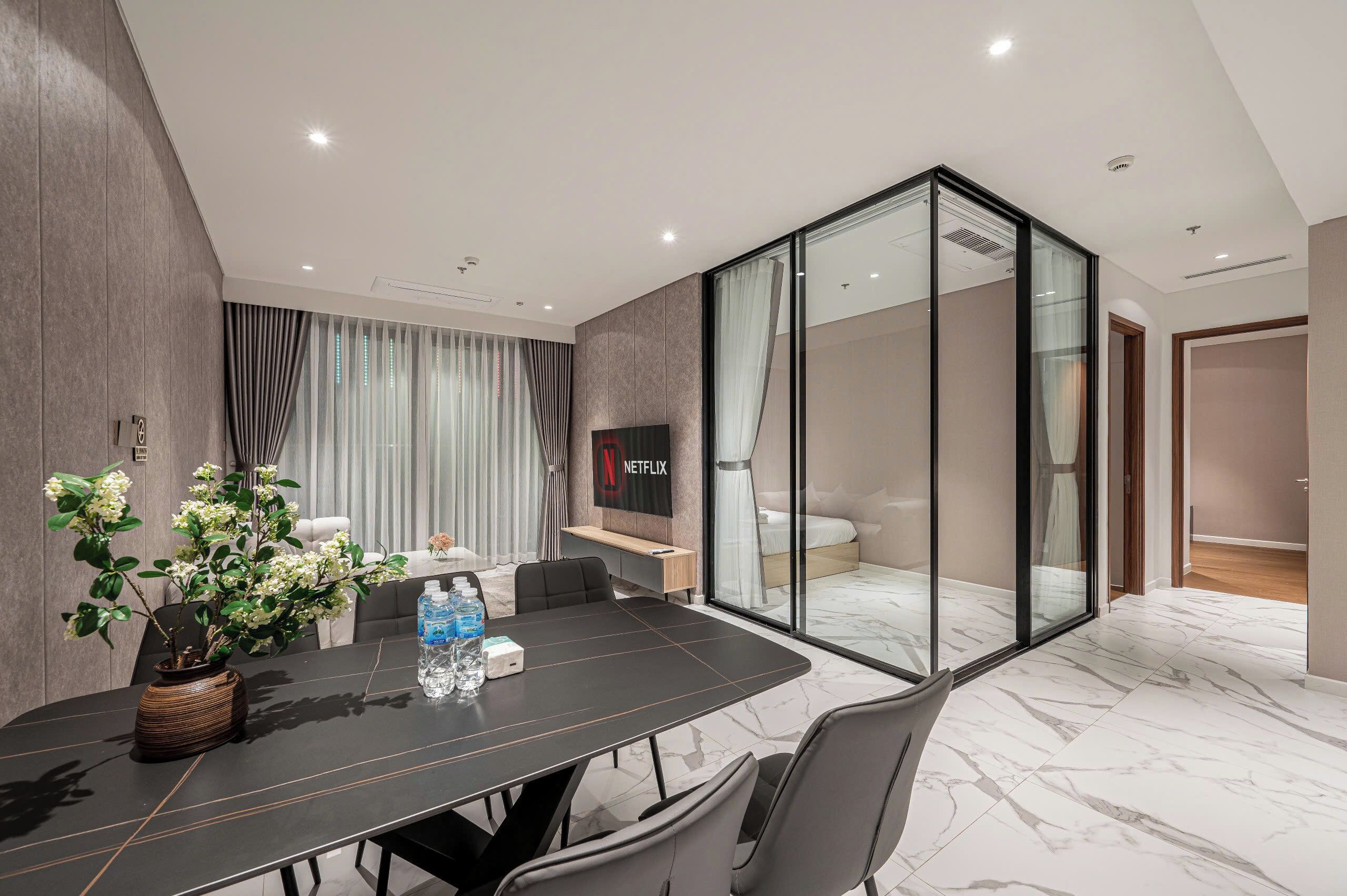 A modern hotel room with a large bed, a glass partition, a flat-screen TV, a dining area with a table and chairs, and a potted plant.