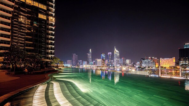 A nighttime cityscape with a river reflecting lights, modern buildings, and a curved waterfront walkway. - Nice Apartments at Metropole Thu Thiem (Ho Chi Minh City)