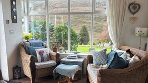 Ein gemütliches Zimmer mit Korbmöbeln, Blick auf eine grüne Landschaft durch große Fenster, und ein kleiner Tisch mit Büchern.
