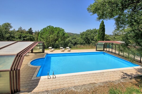 A rectangular swimming pool with a glass enclosure, surrounded by a tiled deck and a glass roof structure.