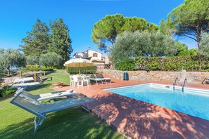 A swimming pool with lounge chairs, a stone wall, and a building with a tiled roof in the background.