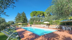 Piscina con sillas de descanso y sombrillas, rodeada de follaje y árboles.
