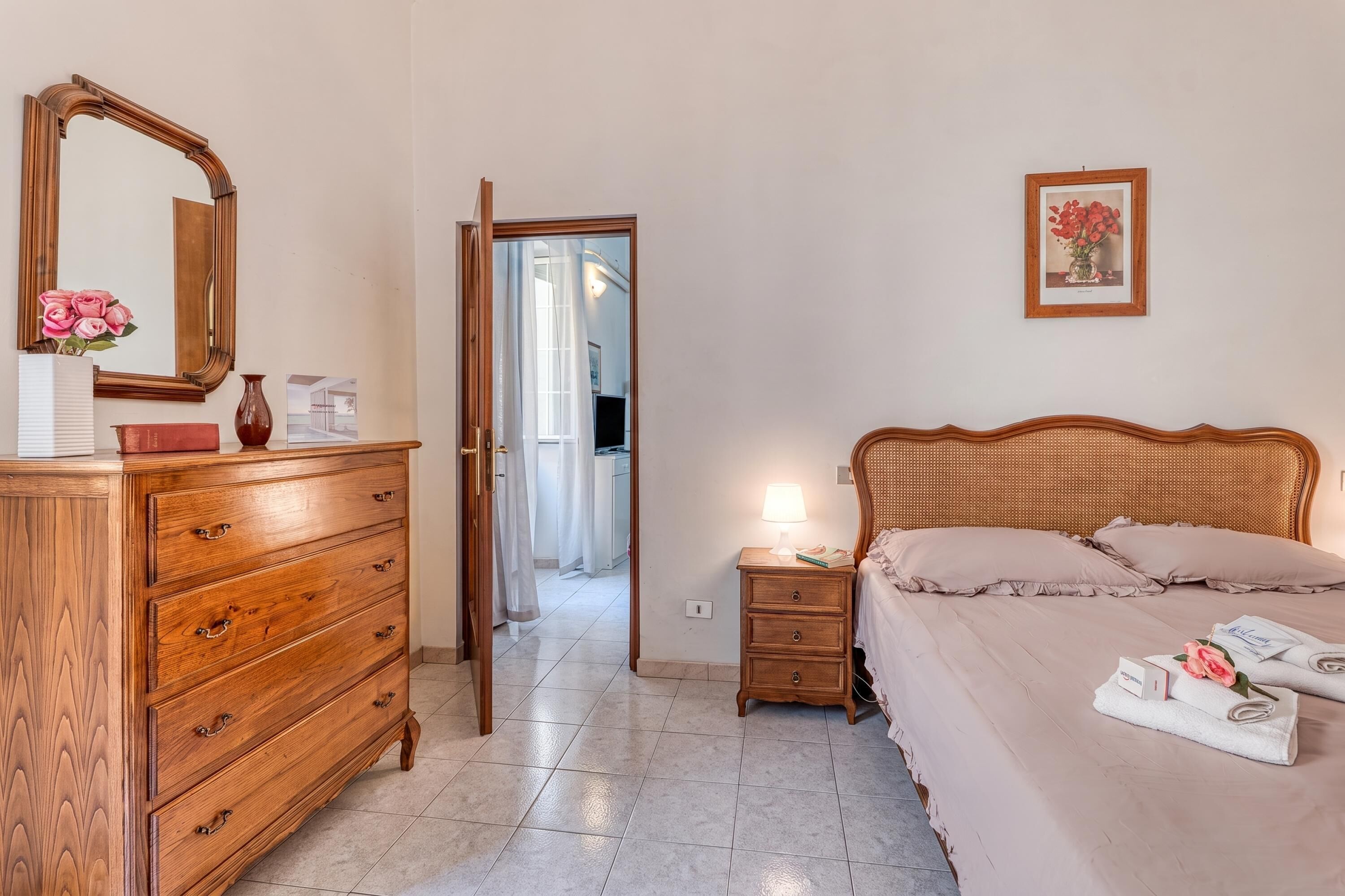 A bedroom with a wooden dresser, a bedside table with a lamp, a bed with pillows and a rolled towel, a framed picture on the wall, and a mirror on the dresser.