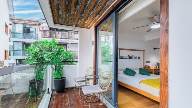 Uma varanda com piso de madeira, plantas em vasos e cadeiras de arame. No interior, há uma cama com cobertor amarelo e um ventilador de teto.