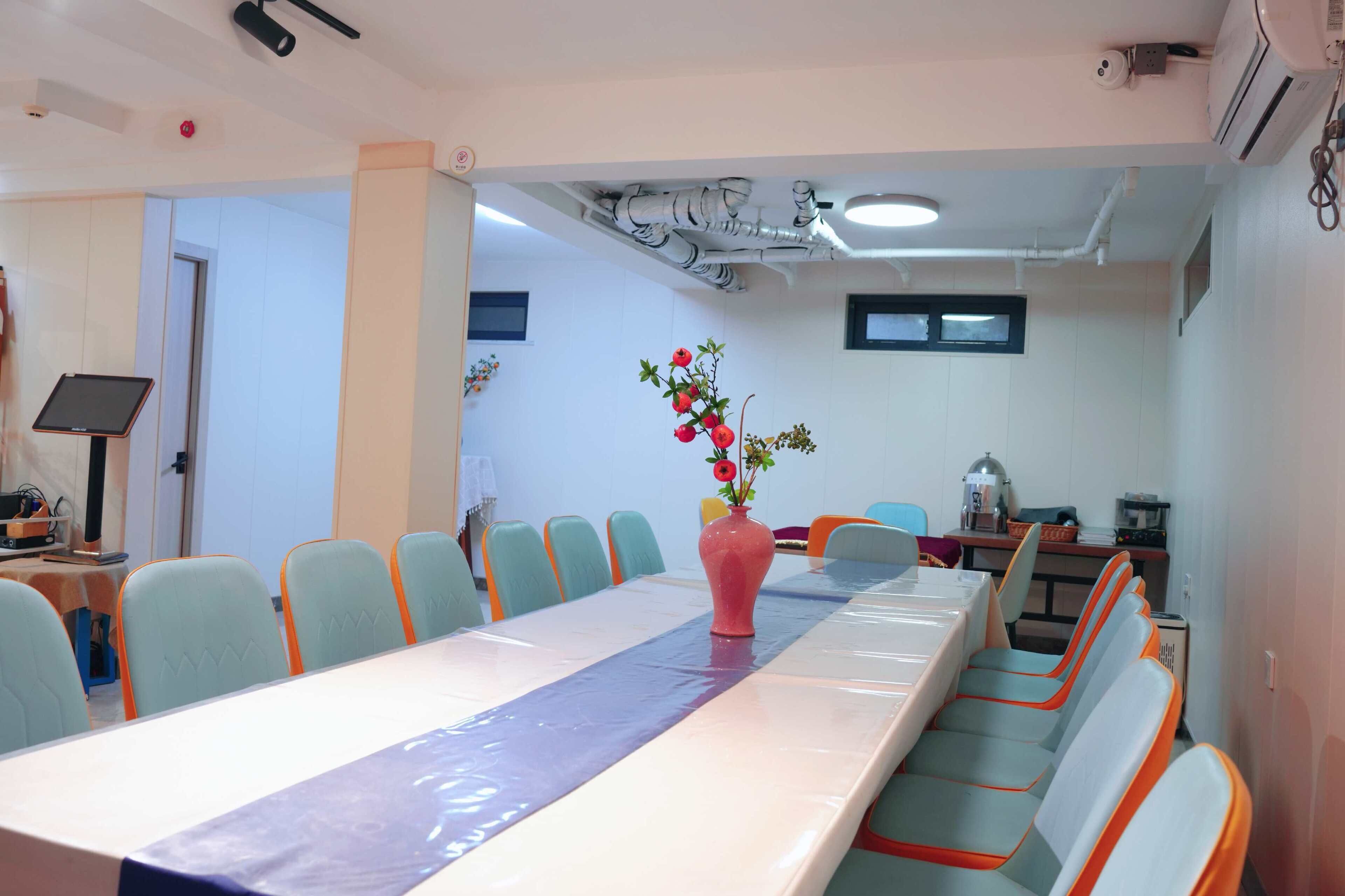 Un salón de conferencias con una larga mesa, sillas, un florero rosa con flores y una pantalla de proyector.