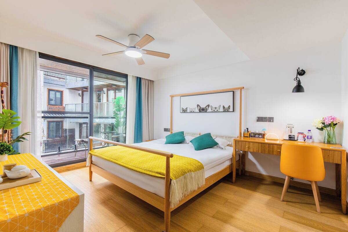 Una habitación de hotel moderna con cama, escritorio y silla, un balcón con vistas y un ventilador de techo.