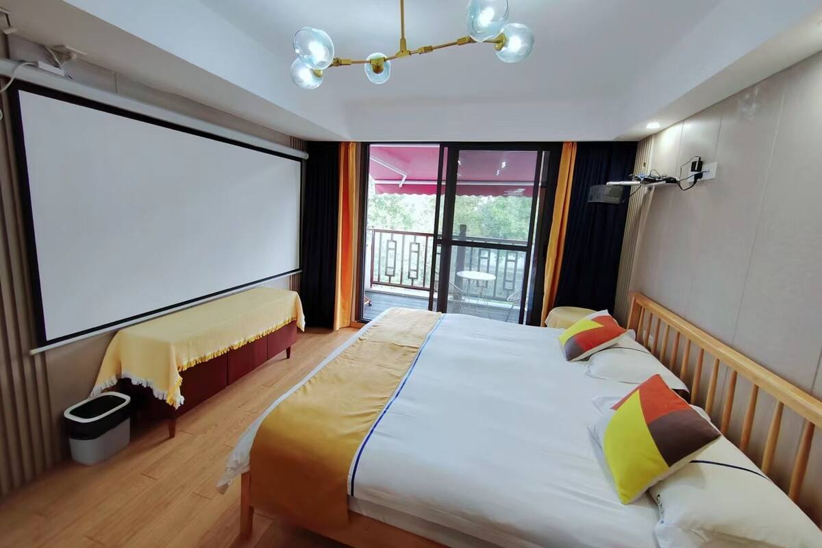 Habitación de hotel con una cama grande, un balcón con mesa y sillas, una lámpara de techo y una pantalla para proyector.