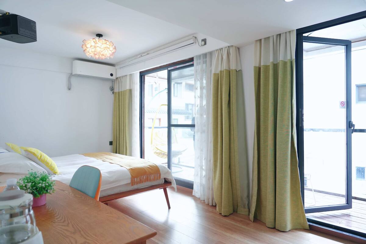 Habitación de hotel con cama, una mesita de madera, una planta en maceta y vistas a través de un ventanal con cortinas.