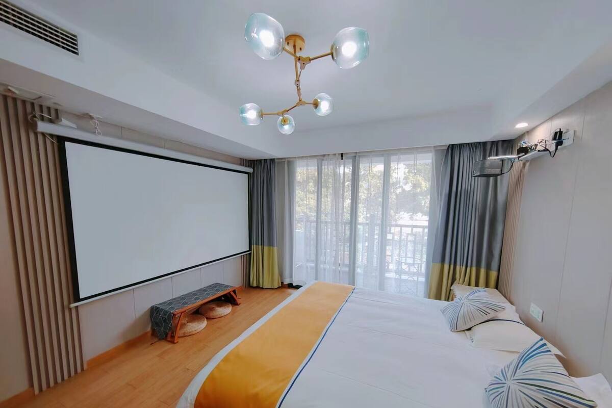 Una habitación de hotel moderna con una cama grande, una lámpara de techo, una pantalla de proyector y vistas al exterior a través de las ventanas.