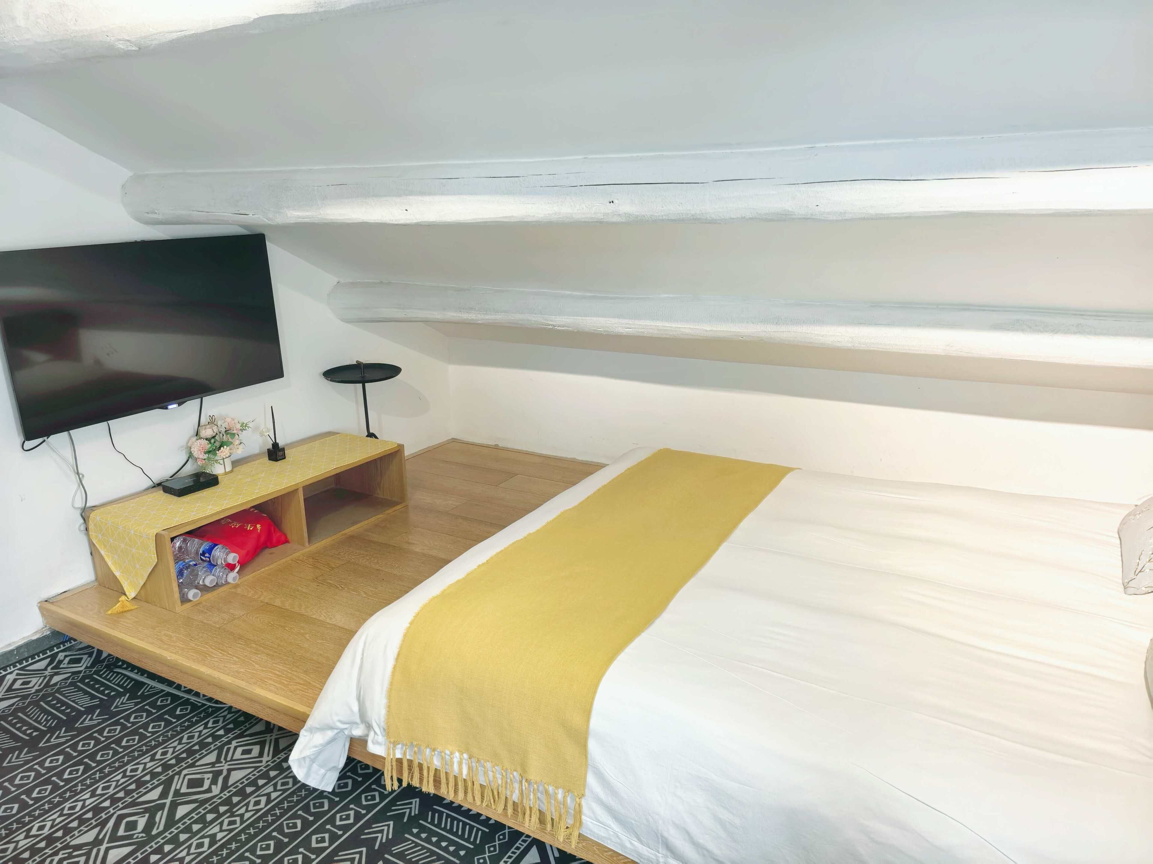 A room with a bed, a TV mounted on the wall, a small wooden table with a yellow cloth, and a patterned rug.