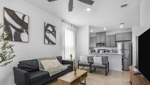 Uma sala de estar moderna com um sofá cinzento, uma mesa de centro de madeira e duas poltronas. Uma cozinha com armários cinzentos e eletrodomésticos de aço inoxidável é visível ao fundo.