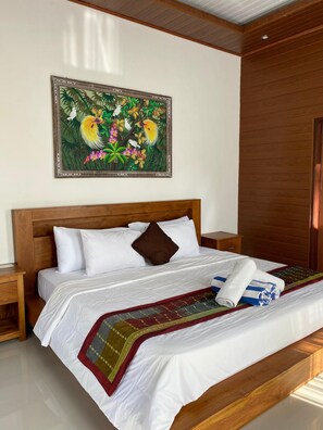 A neatly made bed with white linens and a colorful blanket, a wooden headboard, a nightstand, and a painting of tropical birds and flowers on the wall. - PurnaSari Villa Ubud (Ubud)