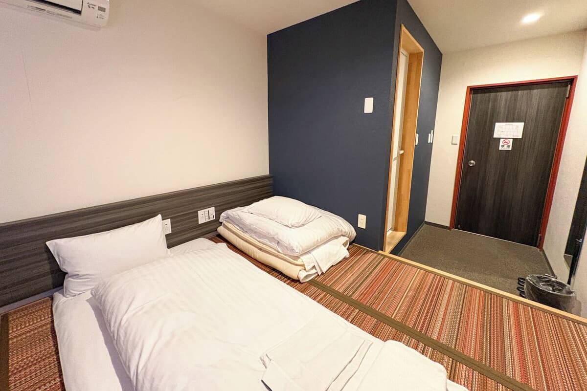 Una habitación de hotel con dos camas, una pared de acento azul oscuro, una puerta y un cubo de basura.