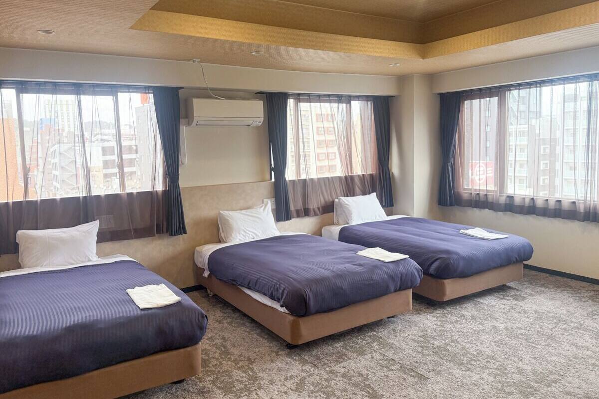 Habitación de hotel con tres camas, un sofá y un ventanal con cortinas.