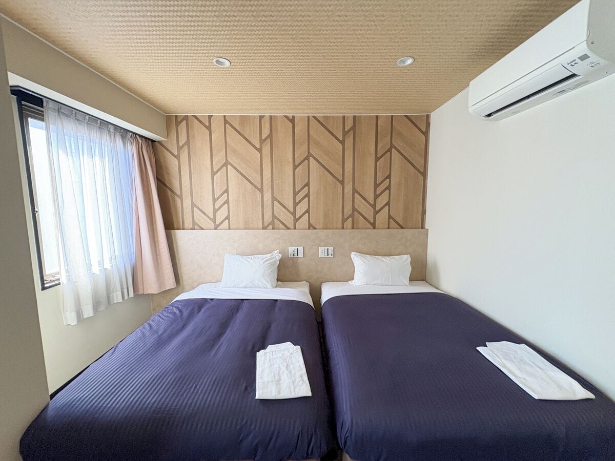 A hotel room with two beds, a wooden headboard, an air conditioning unit, and a window with curtains.