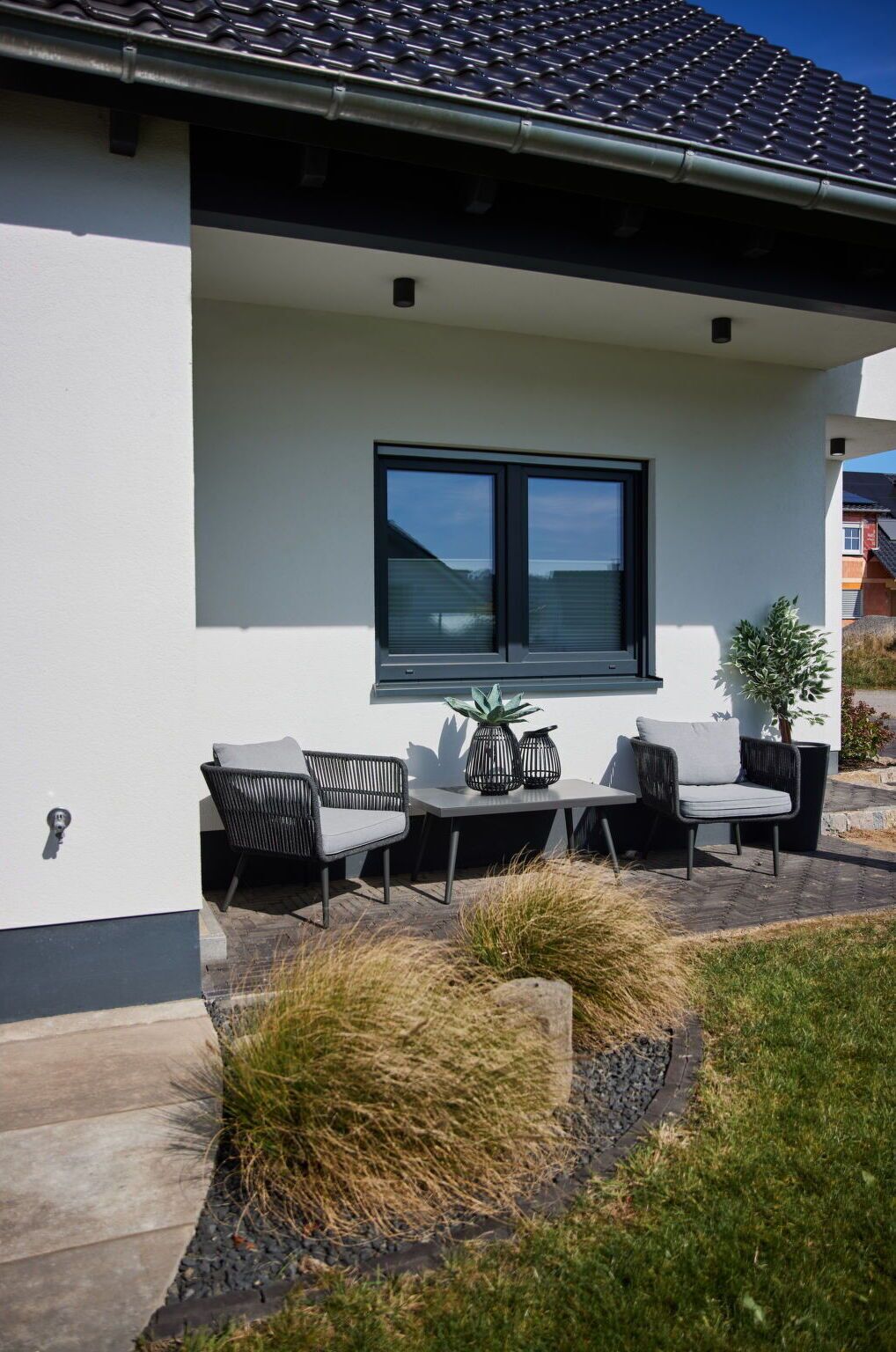 Une maison moderne dotée d’un patio aménagé avec du mobilier d’extérieur, une grande fenêtre et un petit arbre.