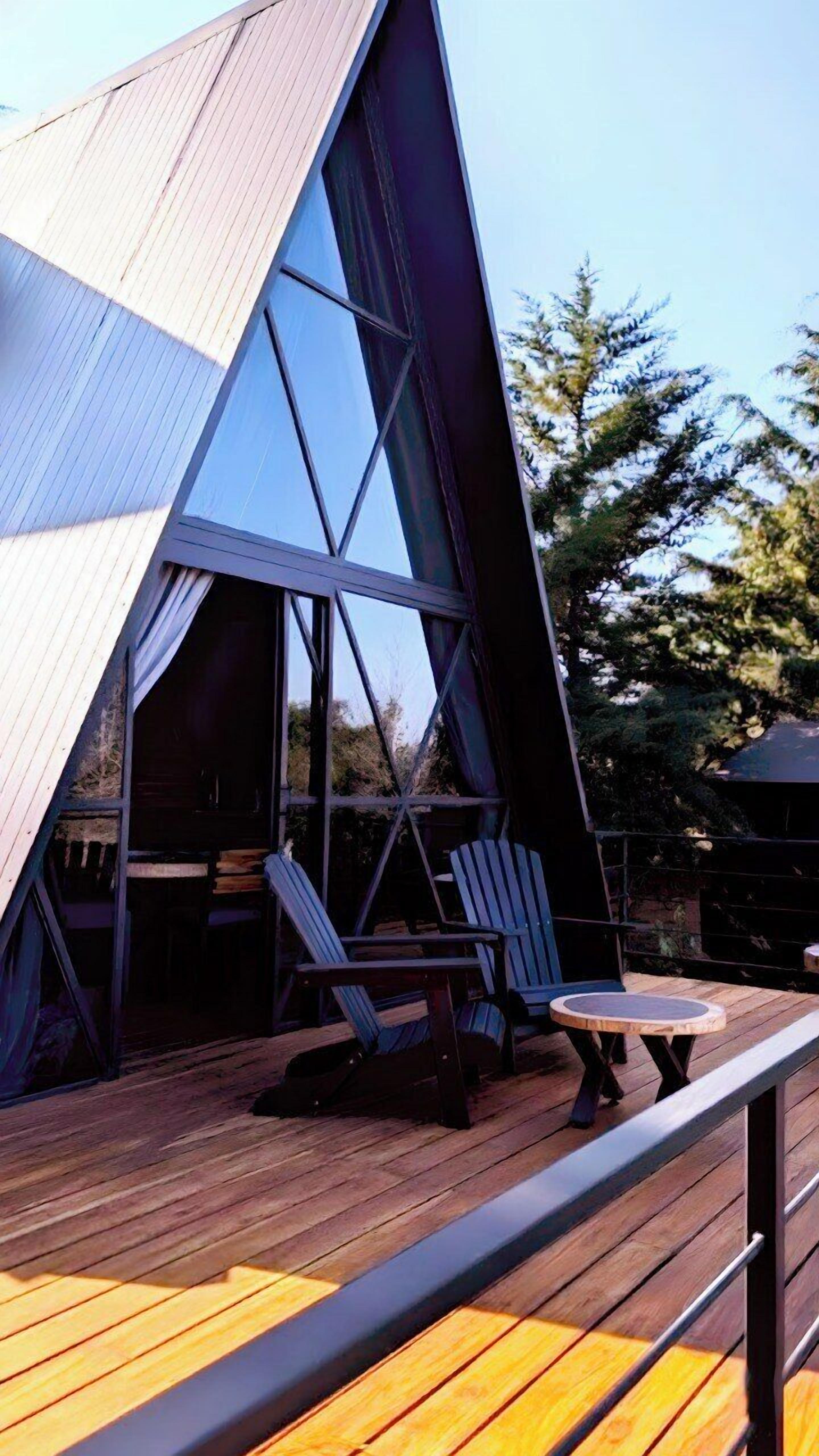 A modern A-frame structure with a wooden deck, two Adirondack chairs, and a small round table.