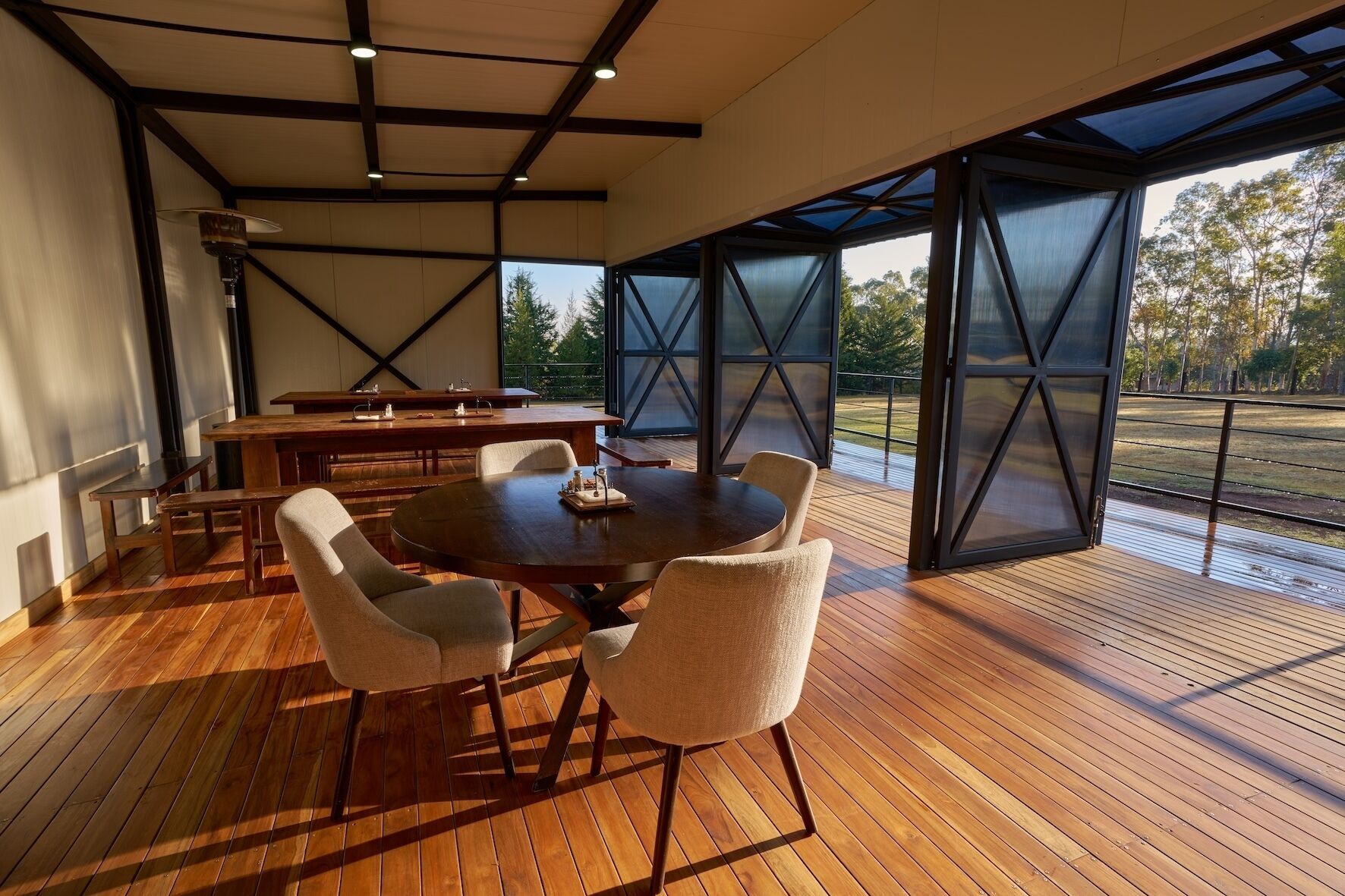 A modern dining area with a round table and chairs, wooden flooring, and large windows offering a view of the outdoors.