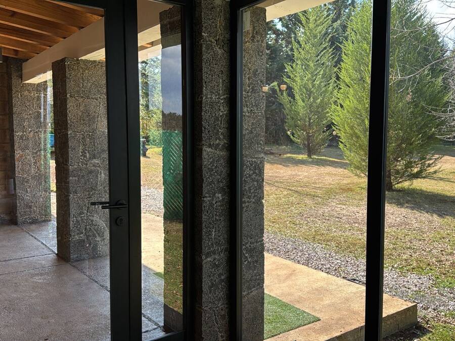 Een overdekt buitengebied met stenen pilaren en een betonvloer, dat uitkomt op een gazon met bomen en een hek.