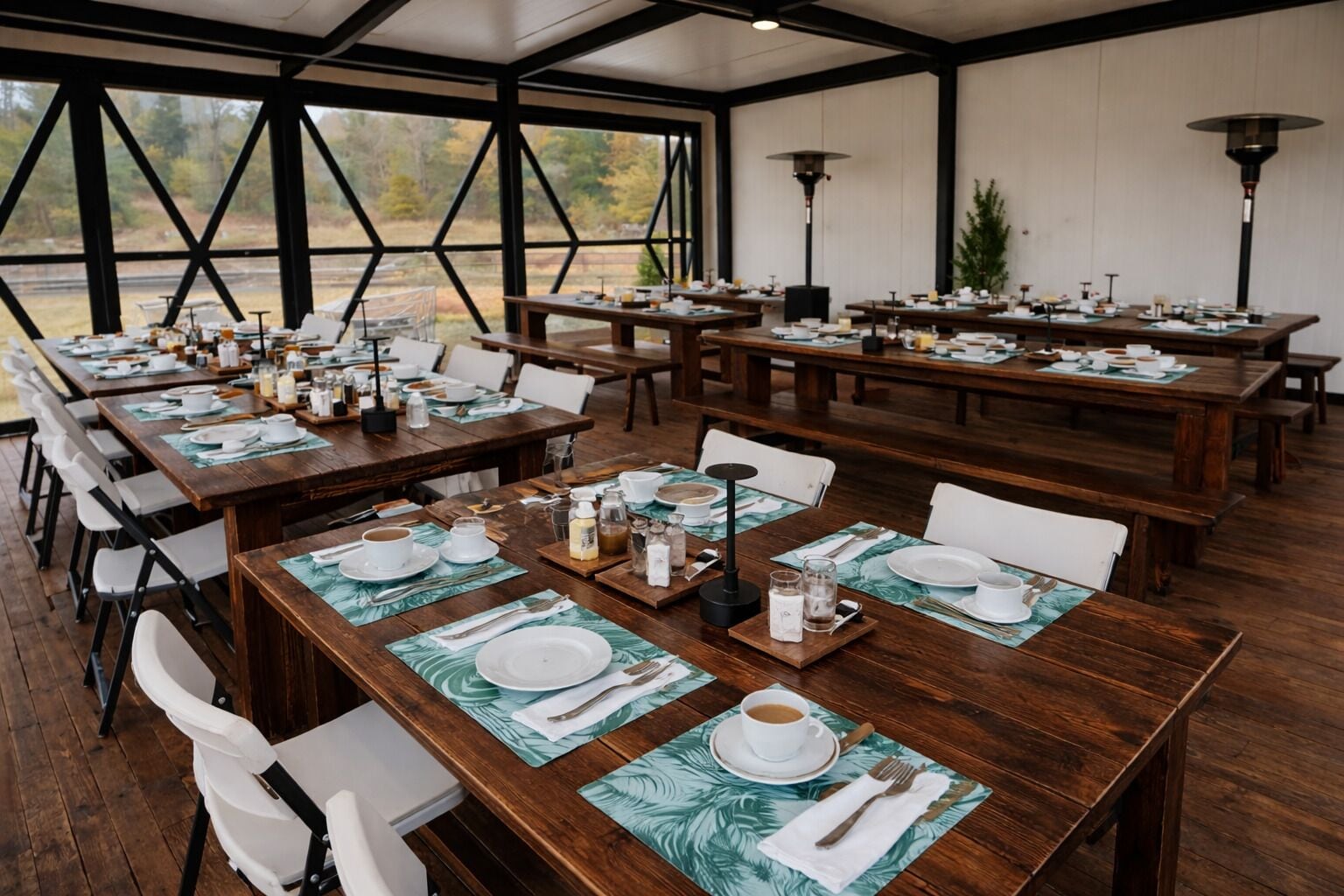 Un comedor amplio con varias mesas de madera preparadas para comidas, sillas blancas y vistas al exterior a través de amplios ventanales.