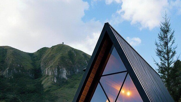 A modern, triangular glass-roofed cabin with a wooden deck, set against a mountainous backdrop.