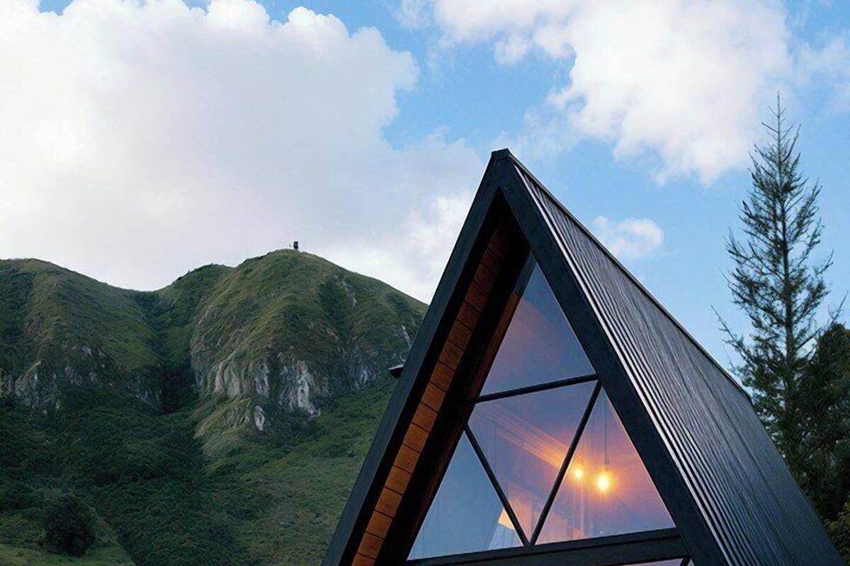 A modern, triangular glass-roofed cabin with a wooden deck, set against a mountainous backdrop.