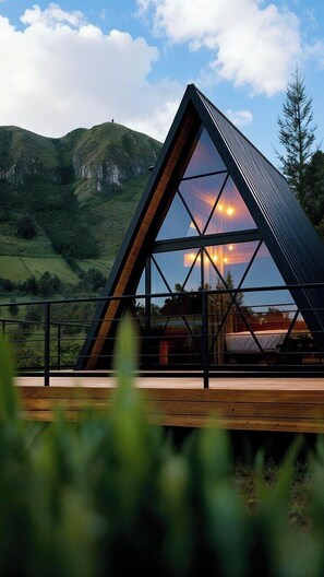Eine moderne, dreieckige Hütte mit Glasdach und einer Holzterrasse, vor einer Berglandschaft gelegen.