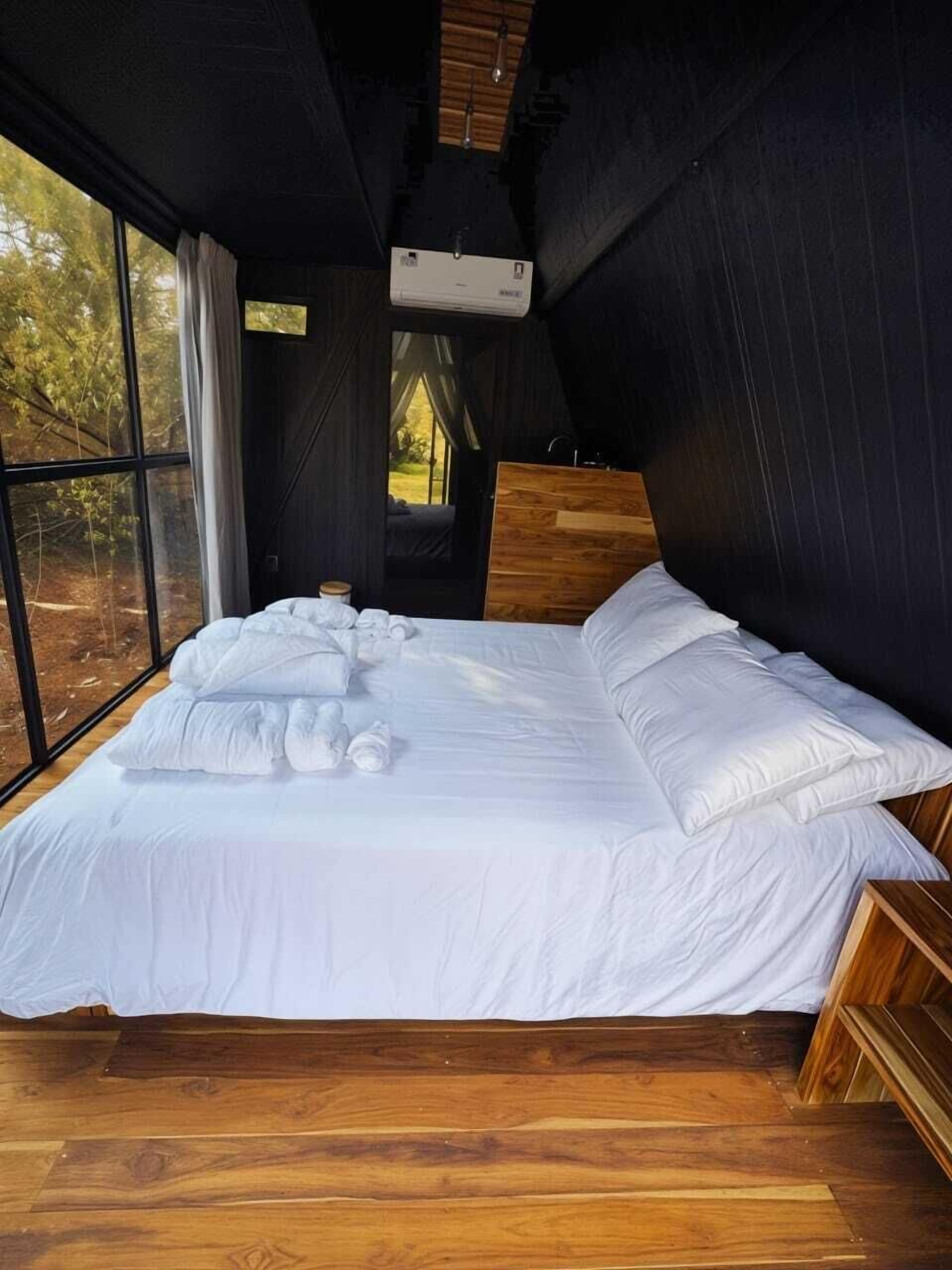 Quarto com uma cama grande, cabeceira de madeira e vista para árvores através de uma janela.