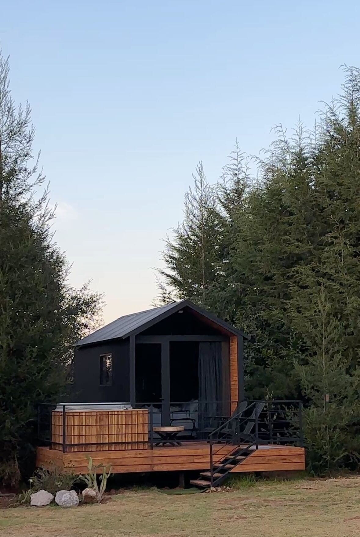 A small cabin with a wooden deck and a metal staircase, surrounded by tall trees.
