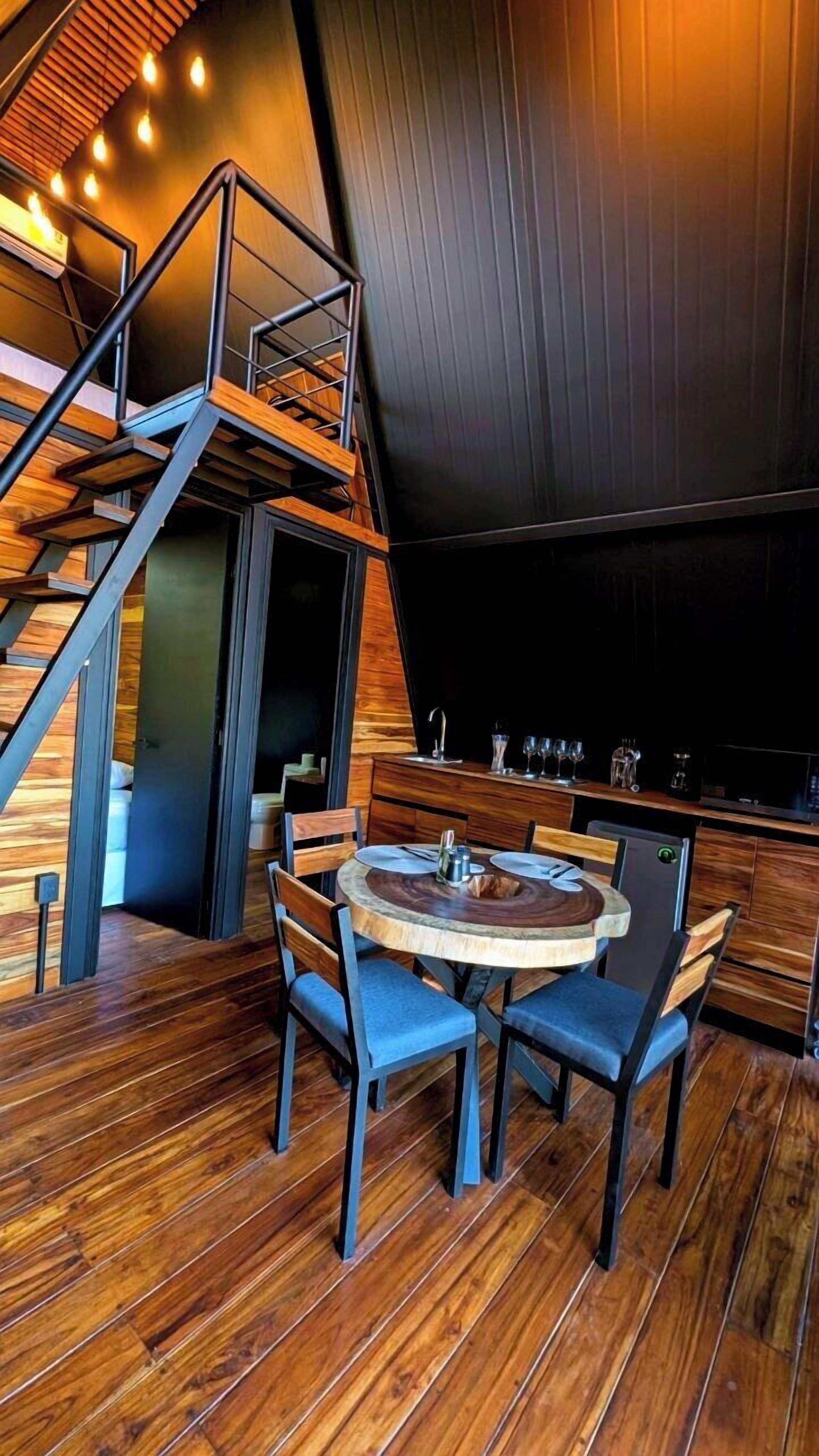 Un interior moderno con una escalera de madera, una mesa redonda de comedor con sillas y una zona de bar con vasos y una cafetera.