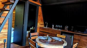 Un interior moderno con una escalera de madera, una mesa redonda de comedor con sillas y una zona de bar con vasos y una cafetera.