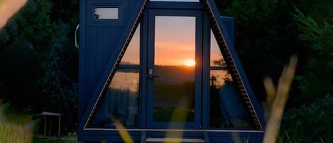Una pequeña cabaña triangular con puerta de cristal, ubicada en una zona cubierta de césped con árboles y montañas al fondo.