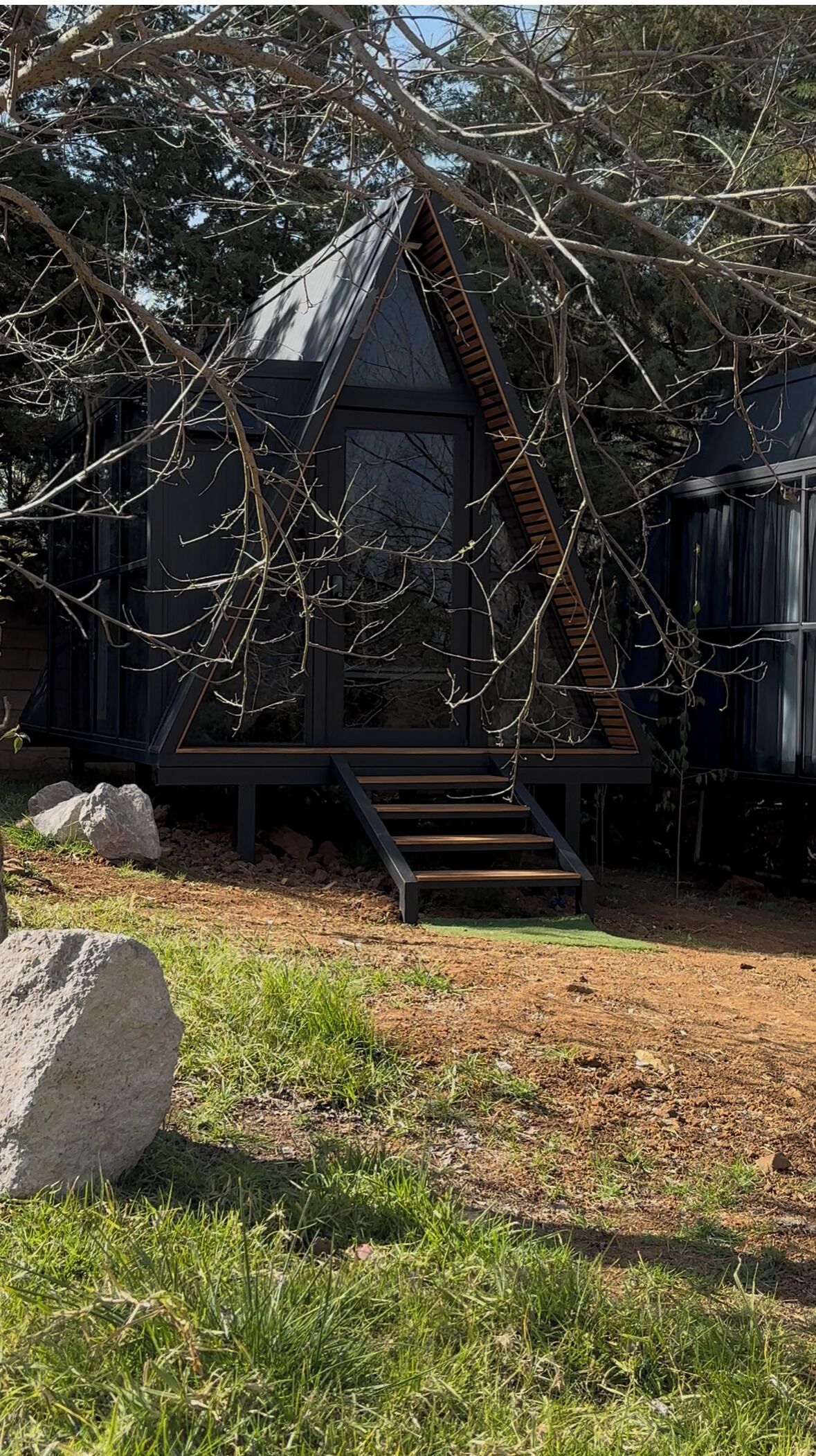 Una moderna baita a forma triangolare con una scala in legno che conduce a un ingresso con porta in vetro, immersa tra alberi e un'area verde.